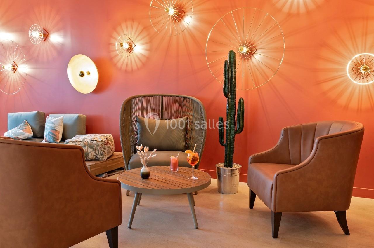 Espace lounge avec fauteuils en cuir, table basse, cactus en pot et mur orange décoré de luminaires design.