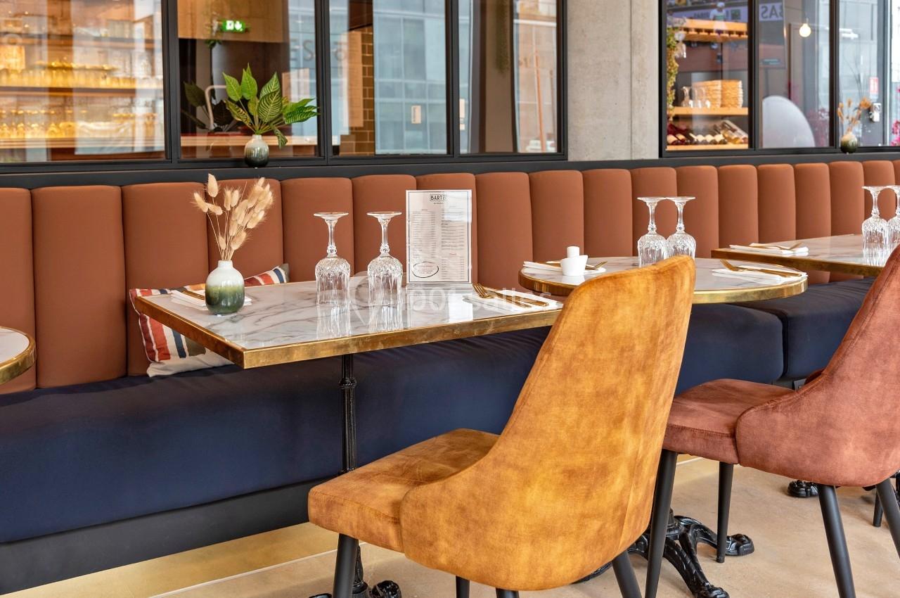 Salle de restaurant avec tables en marbre, chaises rembourrées et banquettes, décorée de plantes et de vaisselle.