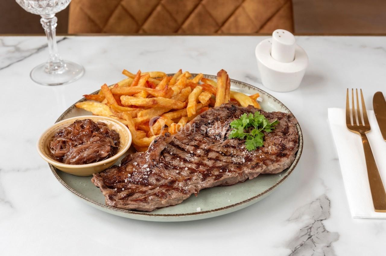 Assiette avec une entrecôte grillée, des frites dorées, une sauce aux oignons et des couverts sur une table en marbre.