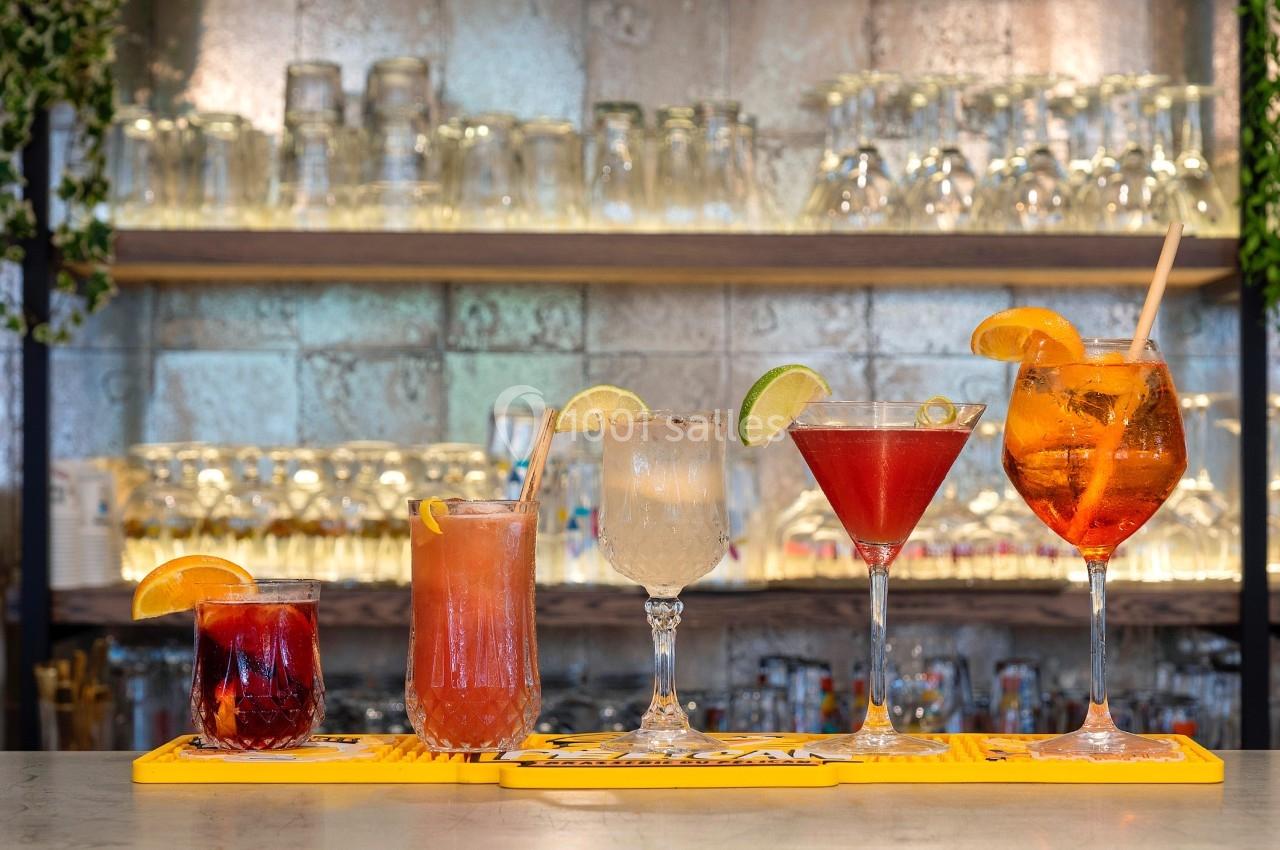 Cinq cocktails colorés alignés sur un bar, décorés de fruits et servis dans des verres variés.