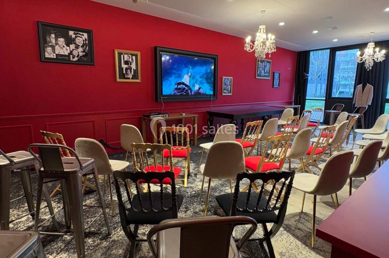 Salle de conférence avec chaises disposées face à un écran, murs rouges décorés de cadres et lustres suspendus.