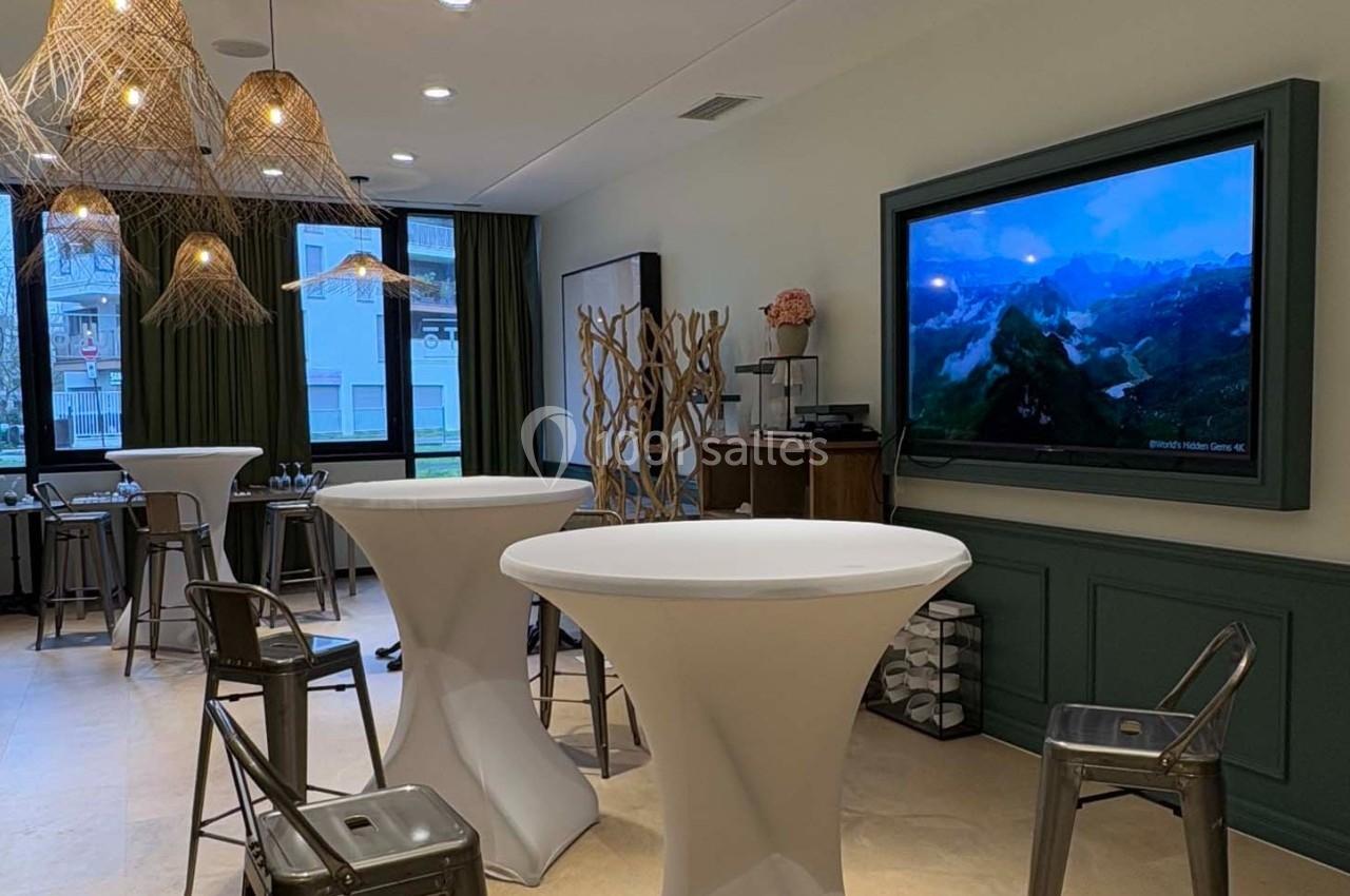 Salle lumineuse aménagée avec des tables hautes, des chaises métalliques et un écran affichant un paysage naturel.