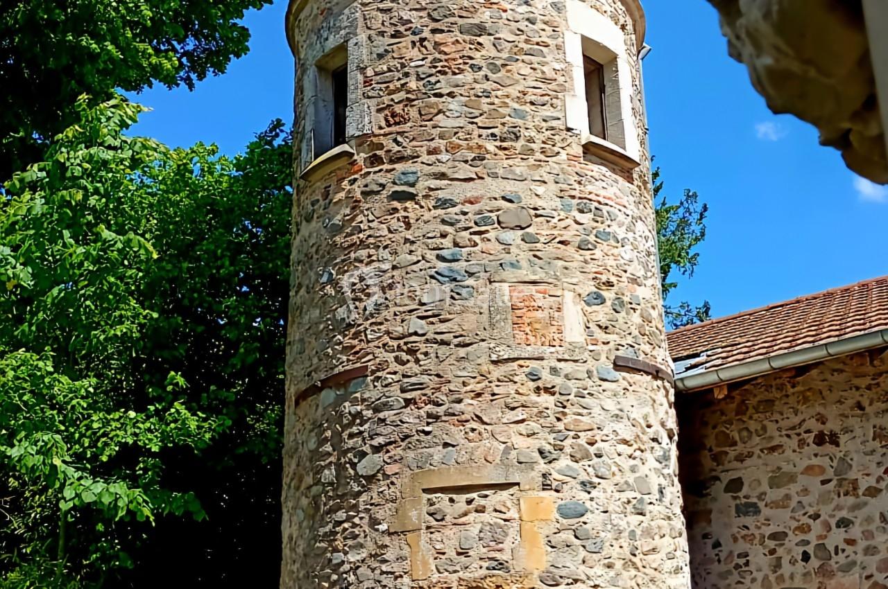 Tour en pierre avec toit conique, entourée de verdure, faisant partie d'un bâtiment ancien sous un ciel bleu.