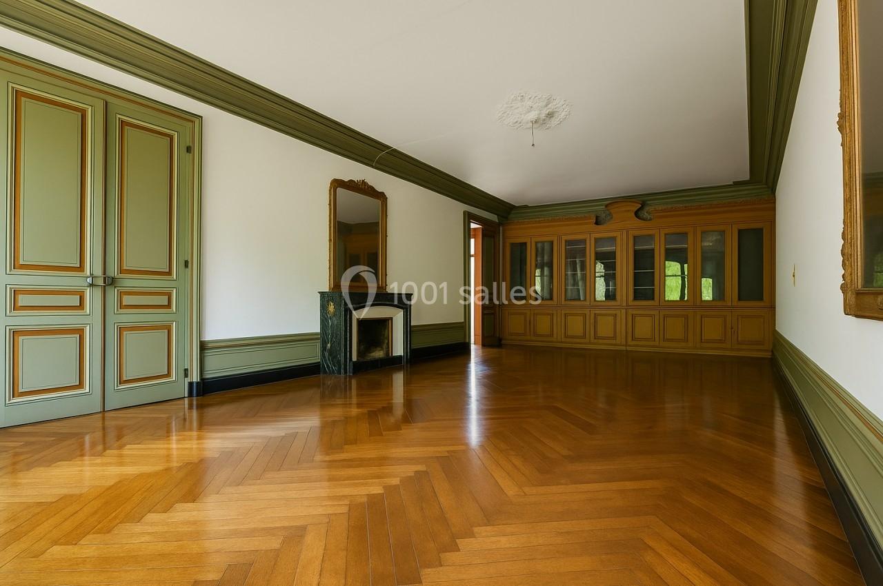 Salle vide avec parquet en bois, cheminée en marbre, miroir, grandes portes vertes et boiseries murales.