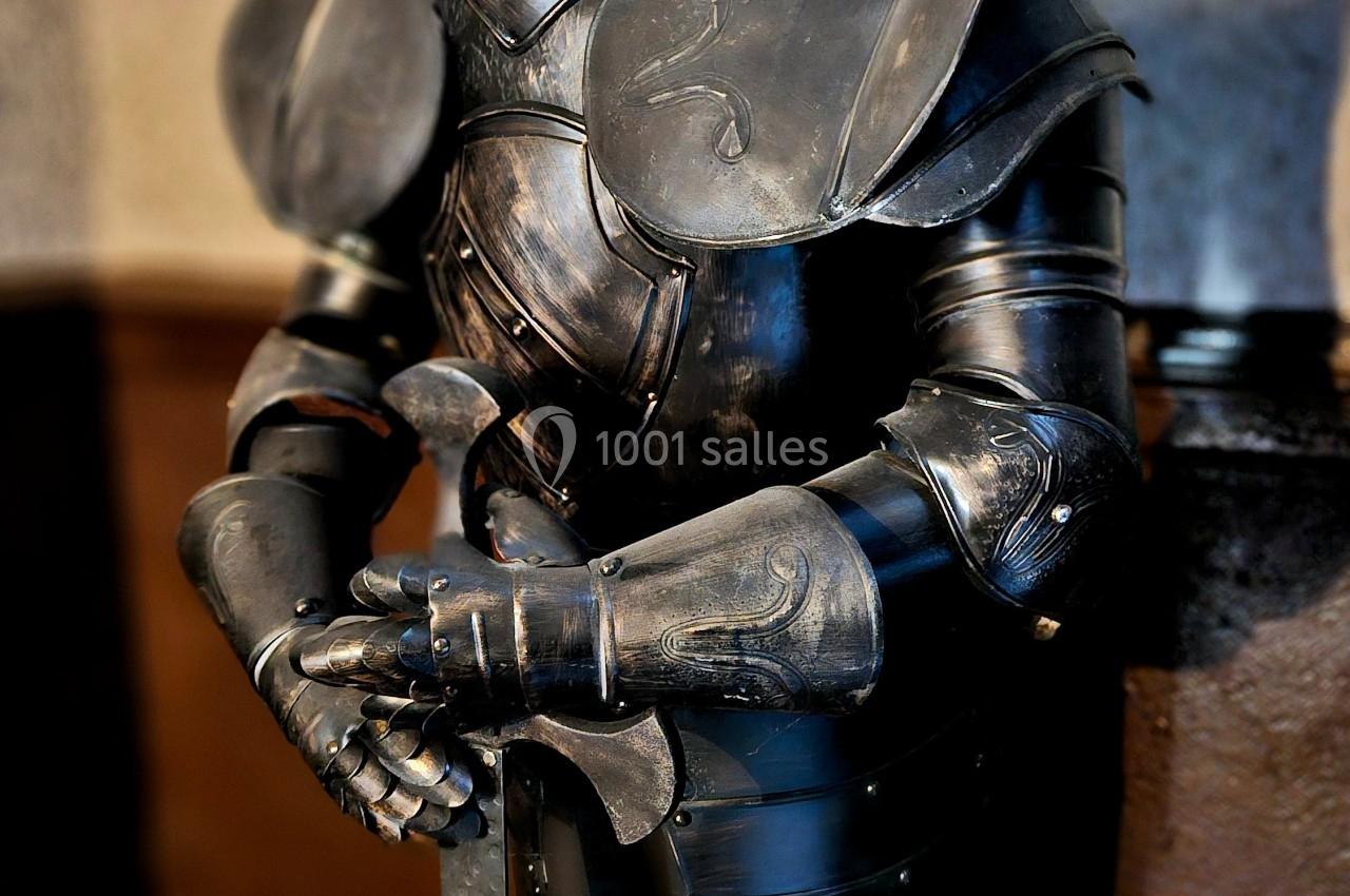 Armure médiévale complète en métal noir, exposée debout dans un intérieur avec un mur texturé en arrière-plan.
