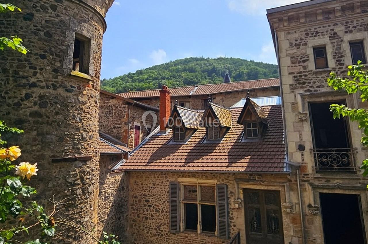Vieilles maisons en pierre avec toits en tuiles, entourées de verdure et d'une colline boisée sous un ciel bleu.