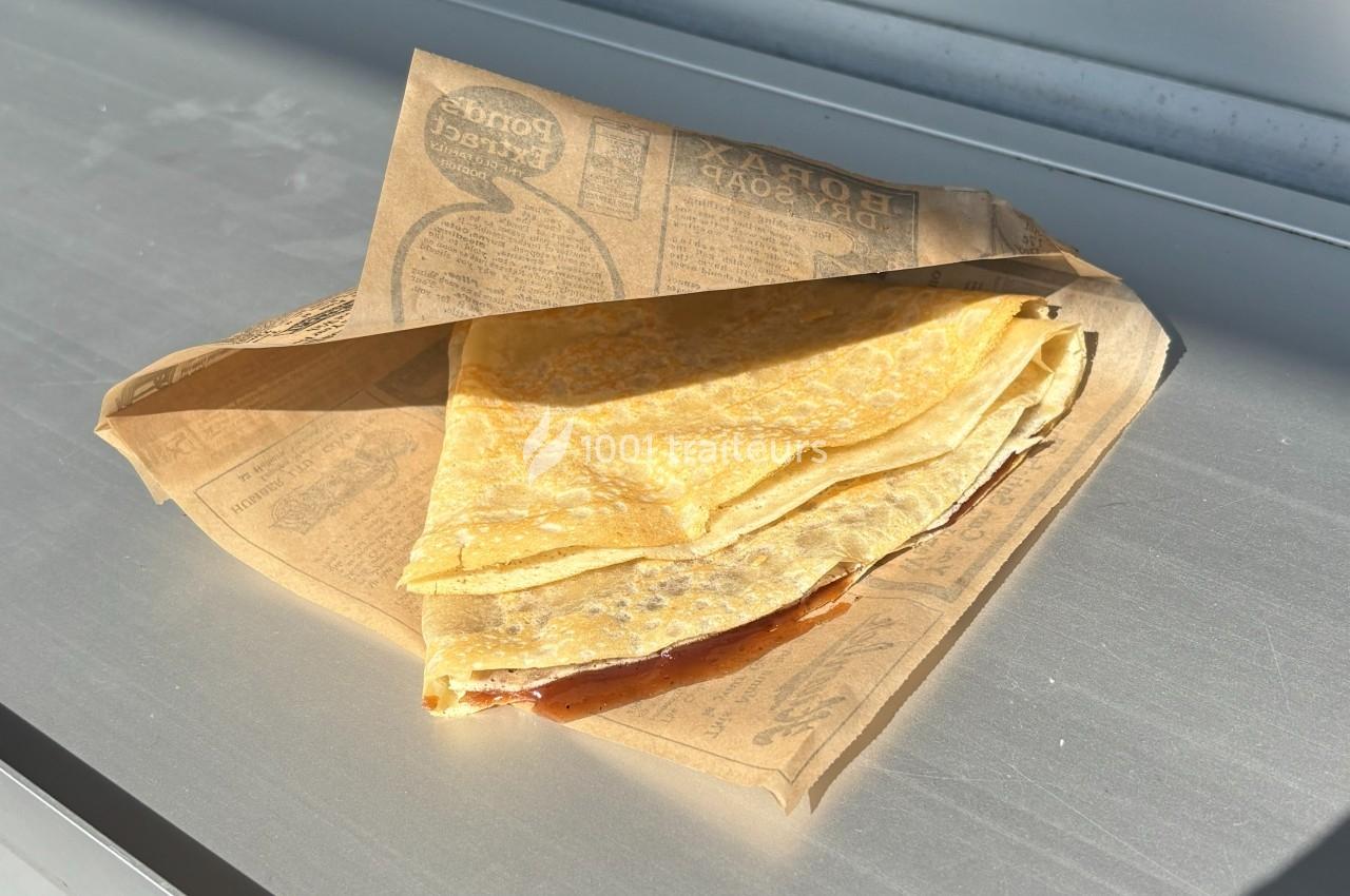 Crêpe pliée dans un papier brun imprimé, posée sur une surface métallique éclairée par la lumière naturelle.