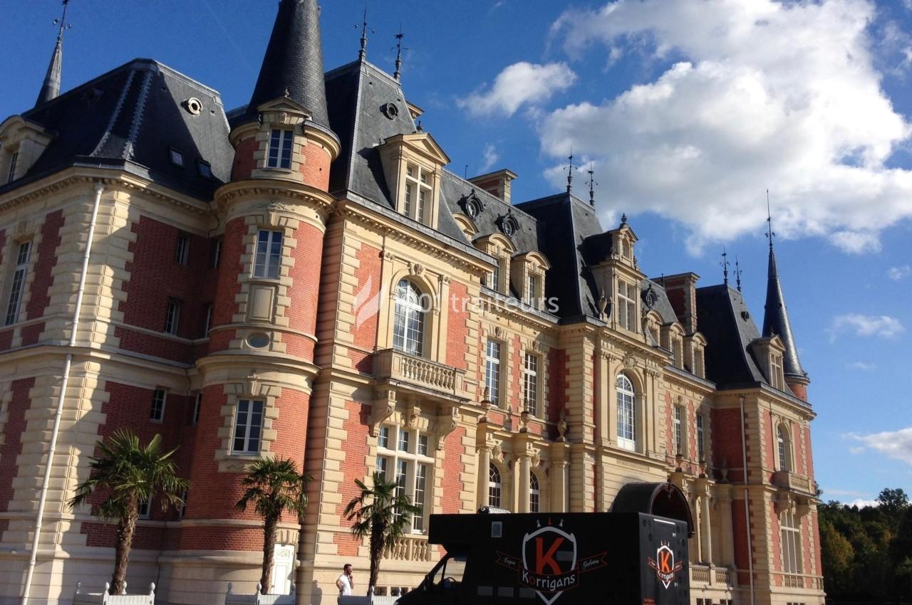 Façade d'un château en briques rouges et pierre claire avec tourelles, sous un ciel bleu partiellement nuageux.