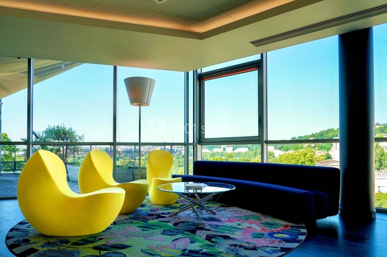 Location salle Caluire-et-Cuire (Rhône) - Le Belvédère #4 Salon moderne avec fauteuils jaunes, canapé bleu, table basse en verre et grande baie vitrée donnant sur un paysage…