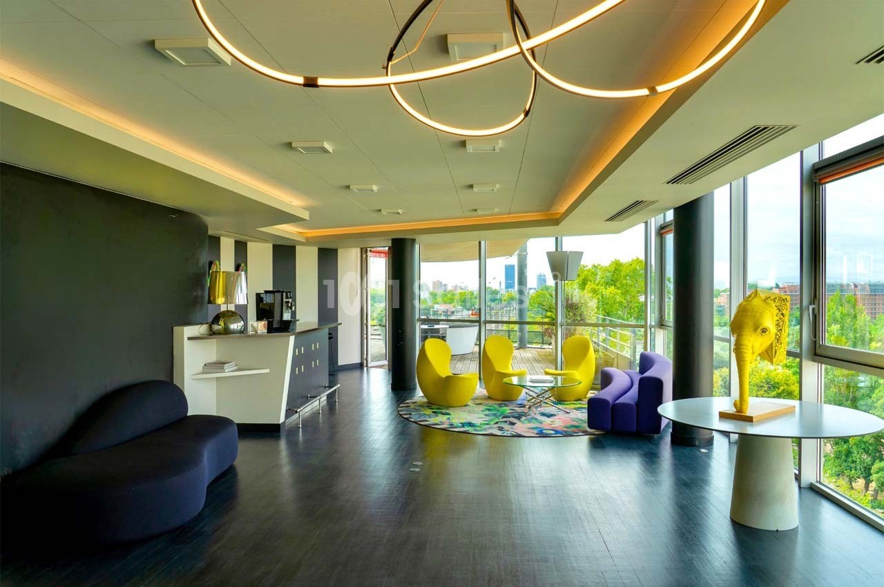 Salle lumineuse avec baies vitrées, mobilier moderne coloré, et vue sur un espace vert urbain.