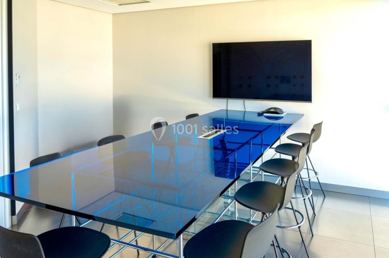 Location salle Caluire-et-Cuire (Rhône) - Le Belvédère #5 Salle de réunion moderne avec une grande table en verre bleu, des chaises noires et un écran mural.