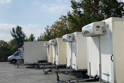 Alignement de remorques frigorifiques blanches stationnées sur un parking, entourées d'arbres et de ciel dégagé.