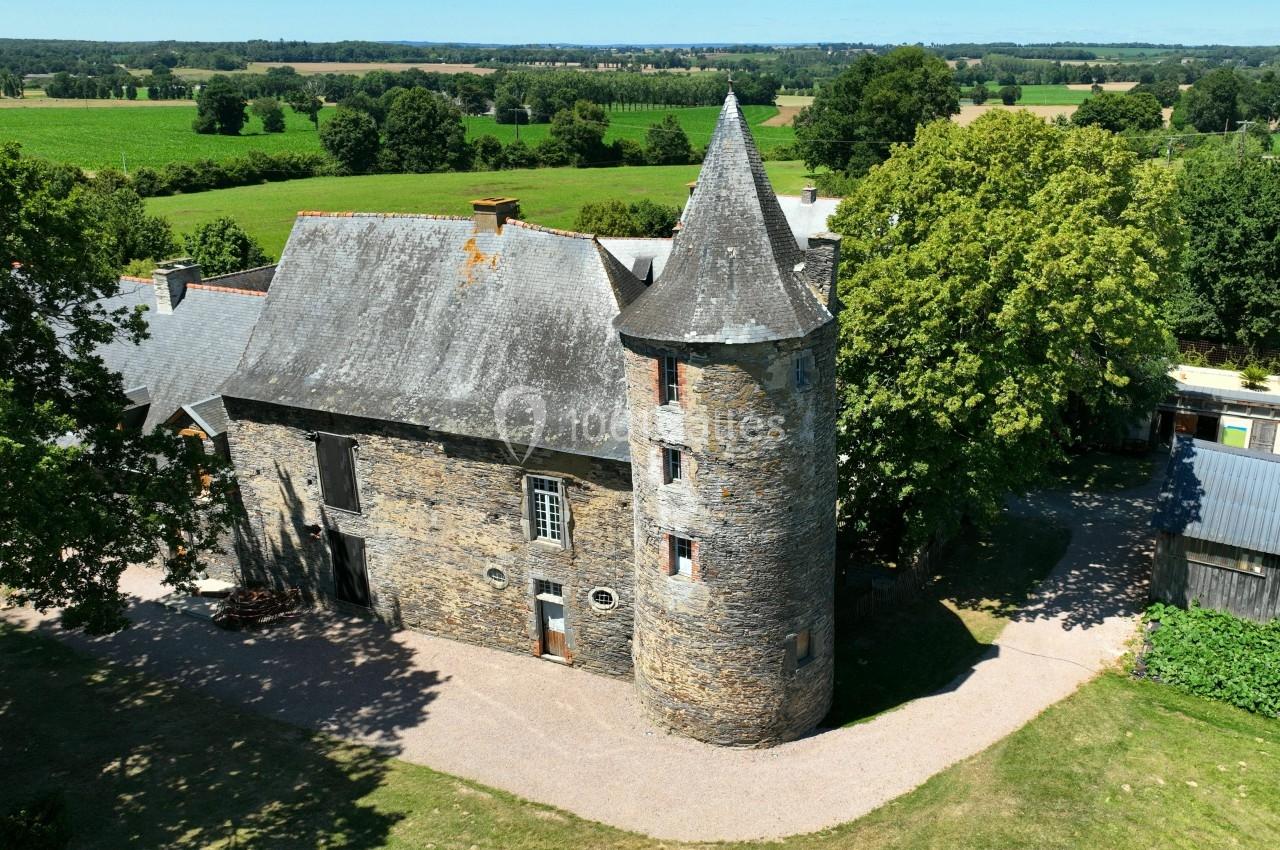 Bâtiment en pierre avec une tour ronde et un toit en ardoise, entouré d'arbres et de champs verdoyants.