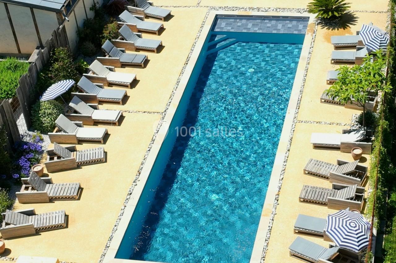 Vue aérienne d'une piscine rectangulaire entourée de transats, parasols et végétation dans un cadre résidentiel.