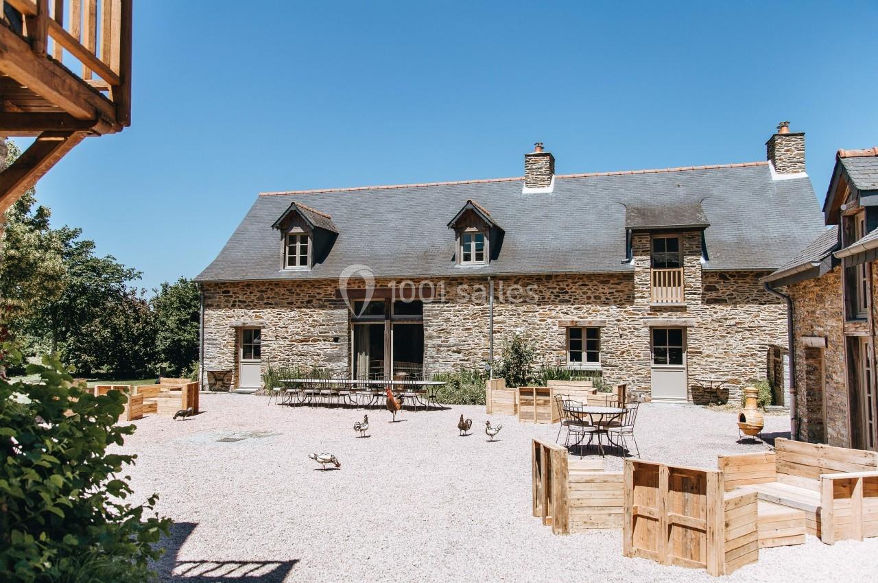 Cour extérieure avec gravier, tables, chaises et bâtiment en pierre avec toit en ardoise sous un ciel bleu.