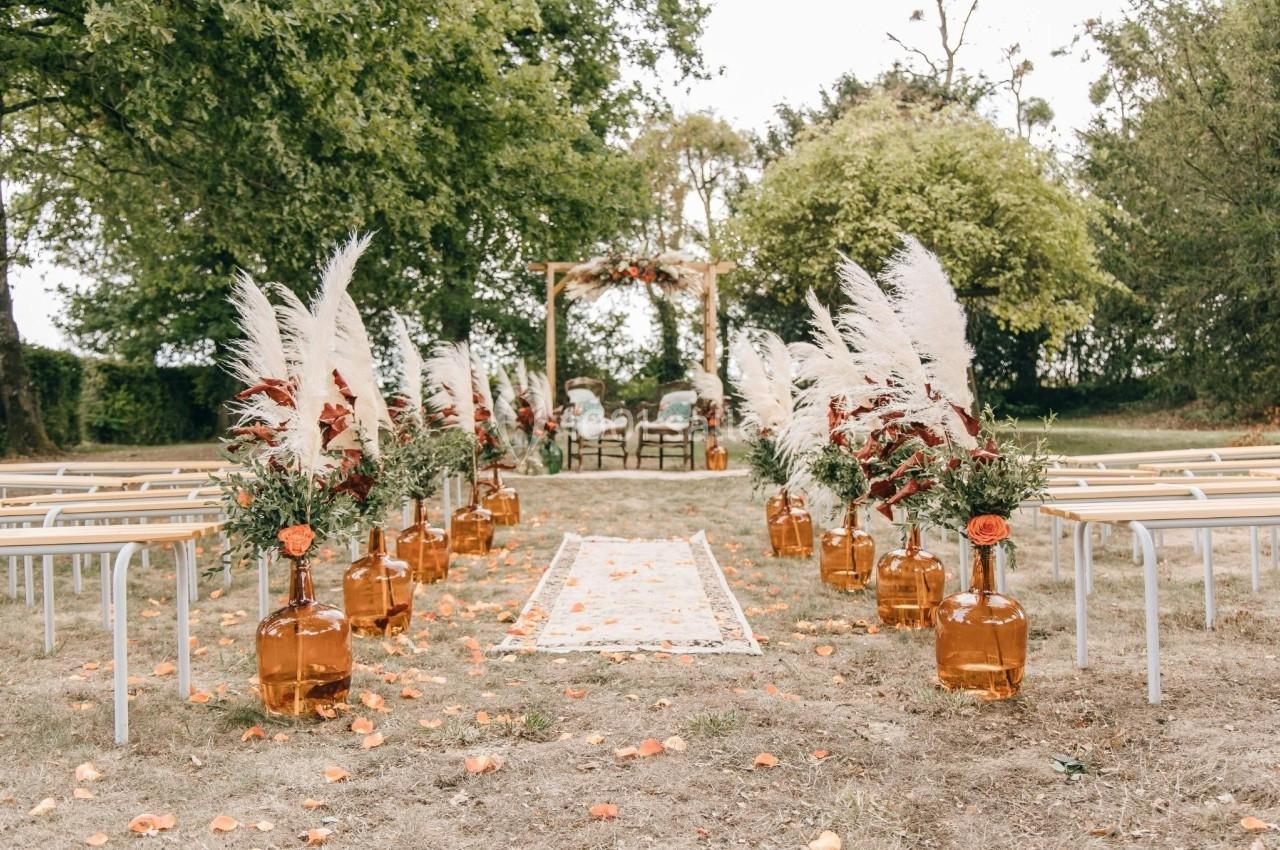 Allée décorée de vases ambrés, pampas et fleurs, menant à une arche en bois dans un jardin pour une cérémonie.