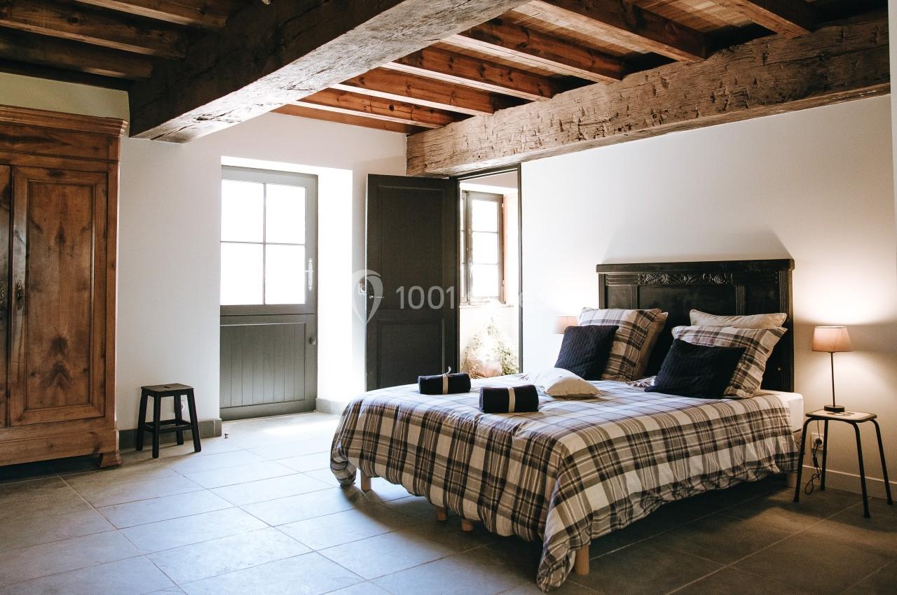 Chambre lumineuse avec lit double, poutres apparentes, armoire en bois, et lumière naturelle entrant par une porte vitrée.