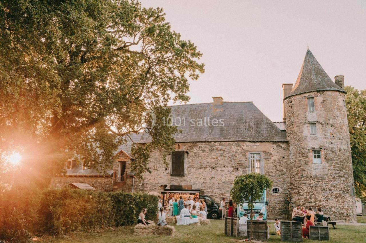 Groupe de personnes rassemblées devant un bâtiment en pierre avec une tour, sous un ciel clair au coucher du soleil.