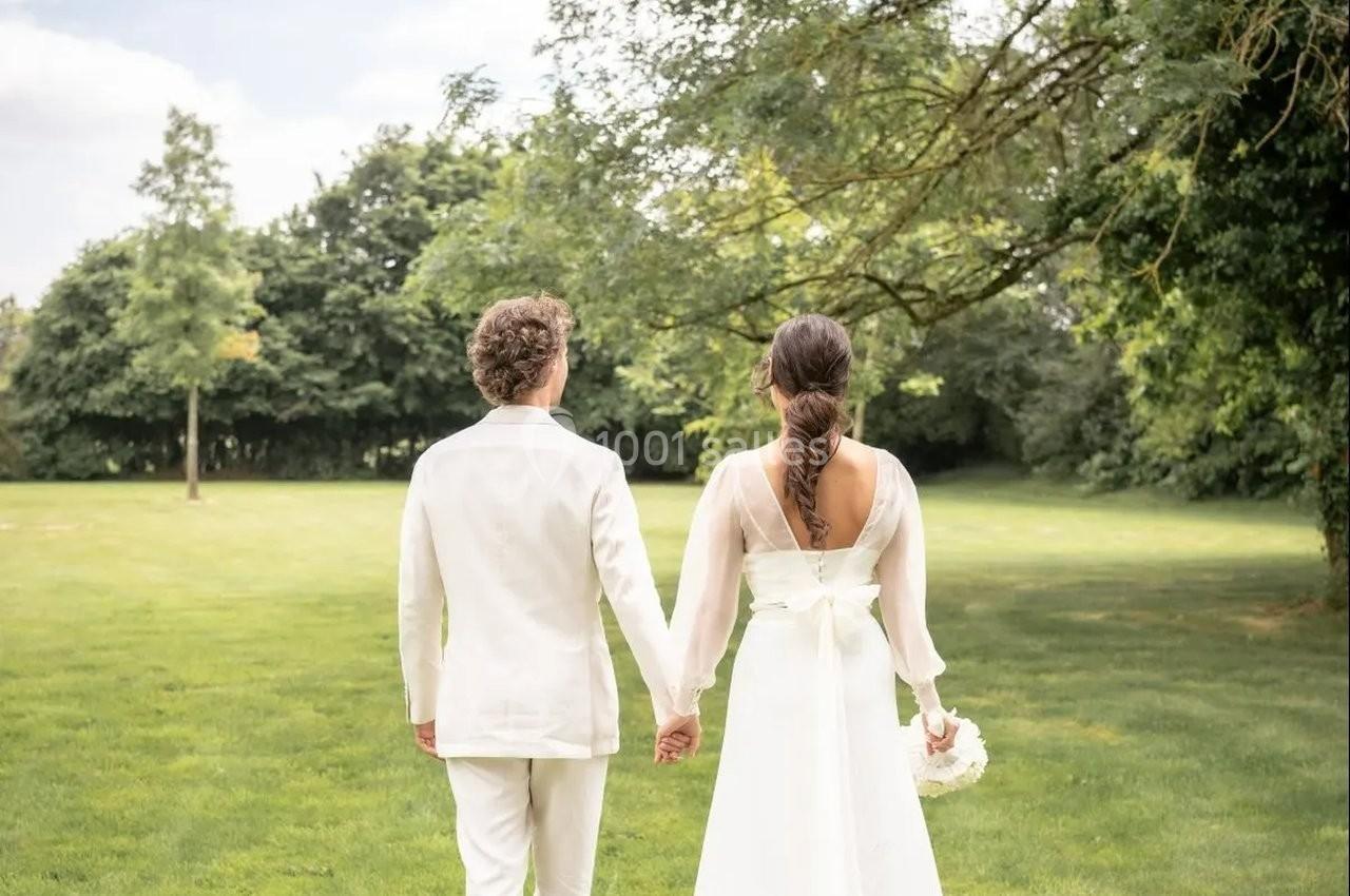 Un couple en tenue de mariage, vu de dos, marchant main dans la main dans un parc verdoyant.