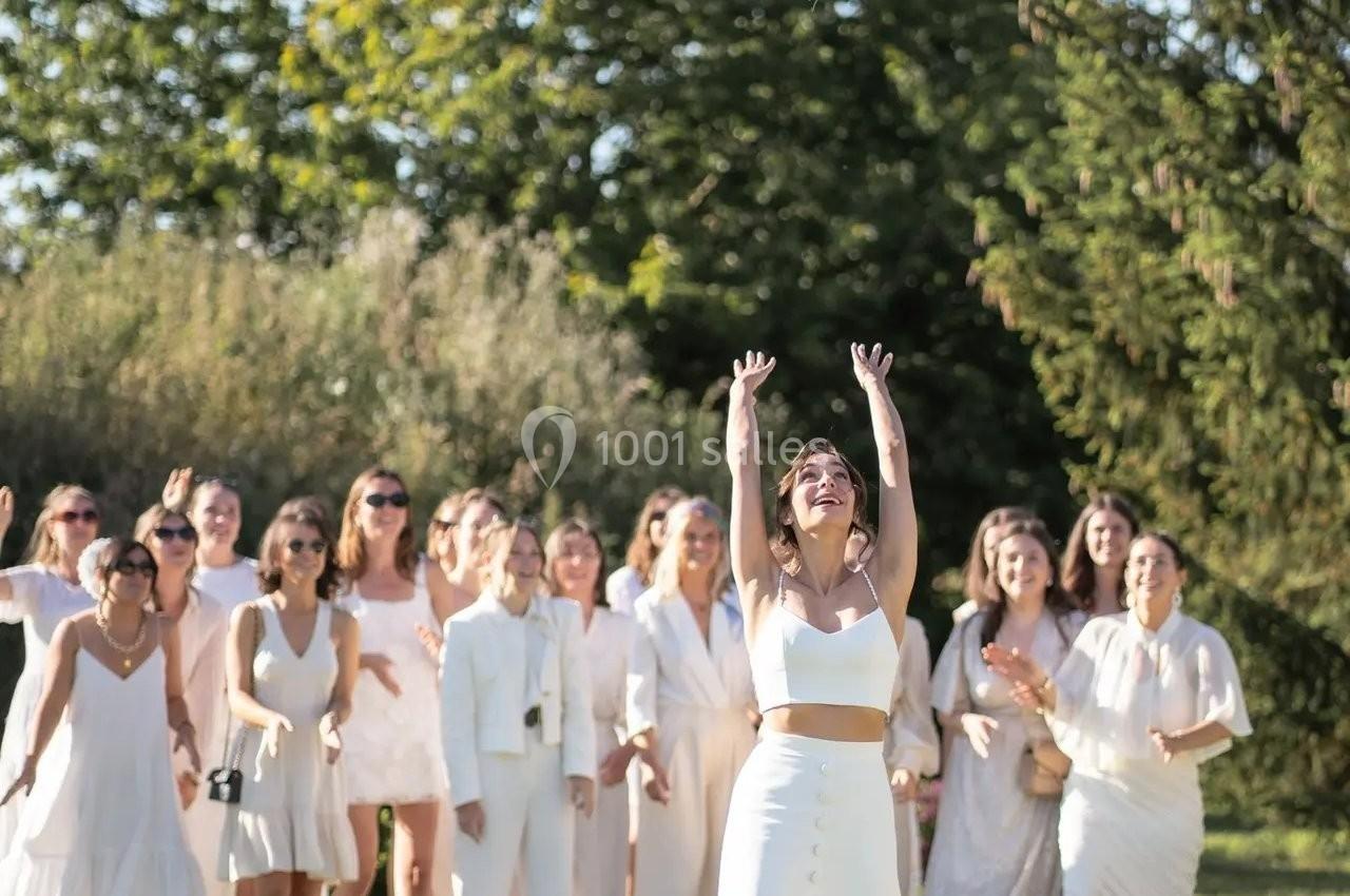 Une mariée lance son bouquet de fleurs vers un groupe de femmes souriantes vêtues de blanc dans un jardin ensoleillé.