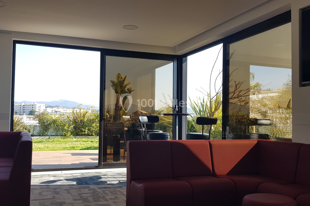Espace intérieur lumineux avec canapés rouges, grandes baies vitrées donnant sur une terrasse et un jardin.