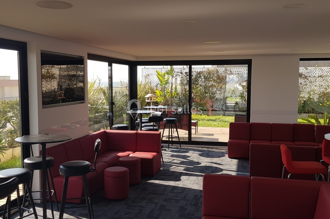 Salle lumineuse avec canapés rouges, tables hautes et vue sur une terrasse végétalisée à travers de grandes baies vitrées.