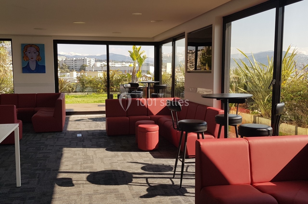 Salle lumineuse avec canapés rouges, tables hautes et grandes baies vitrées donnant sur une terrasse et un jardin.
