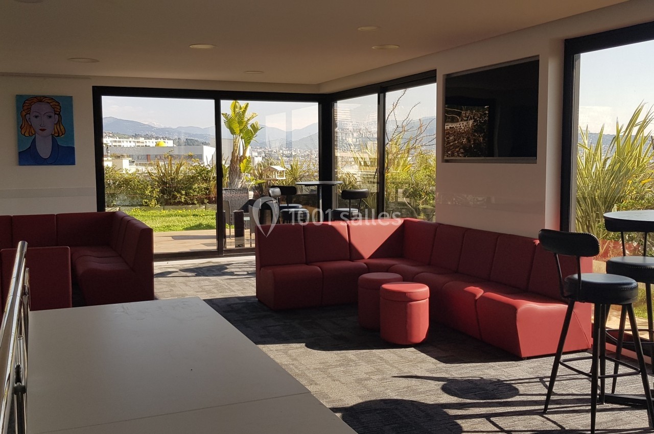Salle lumineuse avec canapés rouges, tables hautes et vue sur une terrasse arborée à travers de grandes baies vitrées.