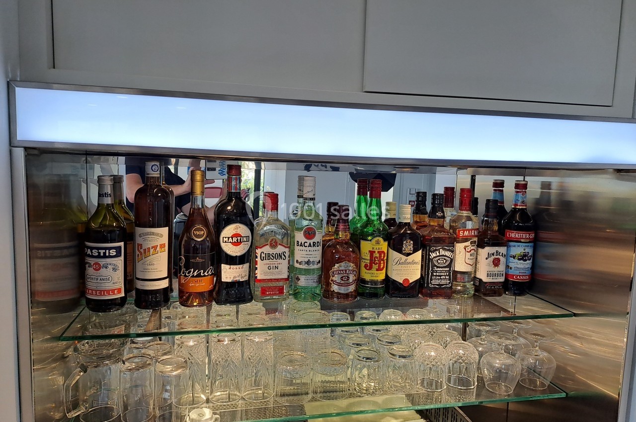 Bouteilles d'alcools variés alignées sur des étagères en verre avec des verres en dessous, dans un meuble éclairé.