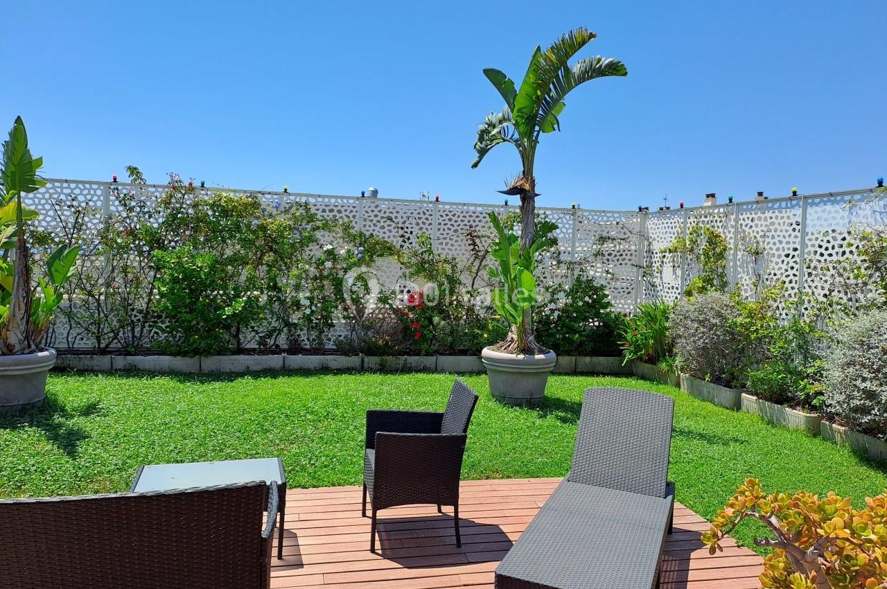 Terrasse en bois avec fauteuils en rotin, donnant sur un jardin verdoyant bordé de plantes et d'un mur ajouré.