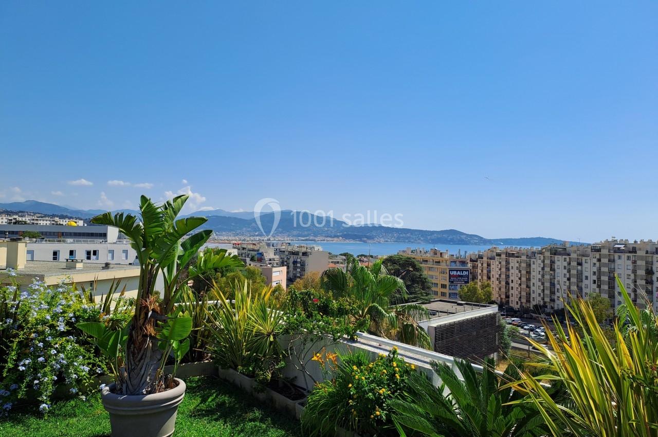 Vue depuis une terrasse avec des plantes en pot, donnant sur des immeubles, des collines et la mer sous un ciel dégagé.