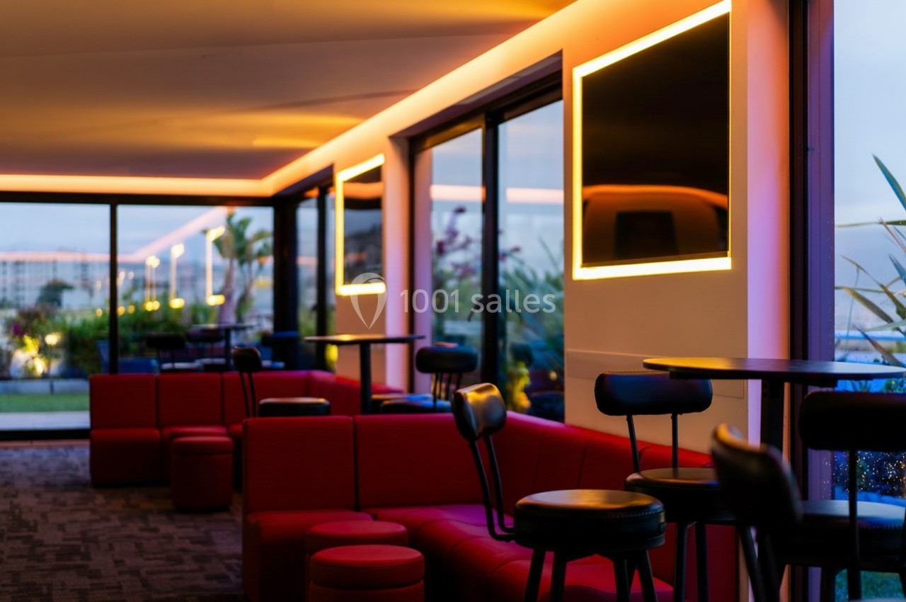 Espace intérieur moderne avec banquettes rouges, tables hautes et grandes baies vitrées donnant sur un extérieur éclairé.