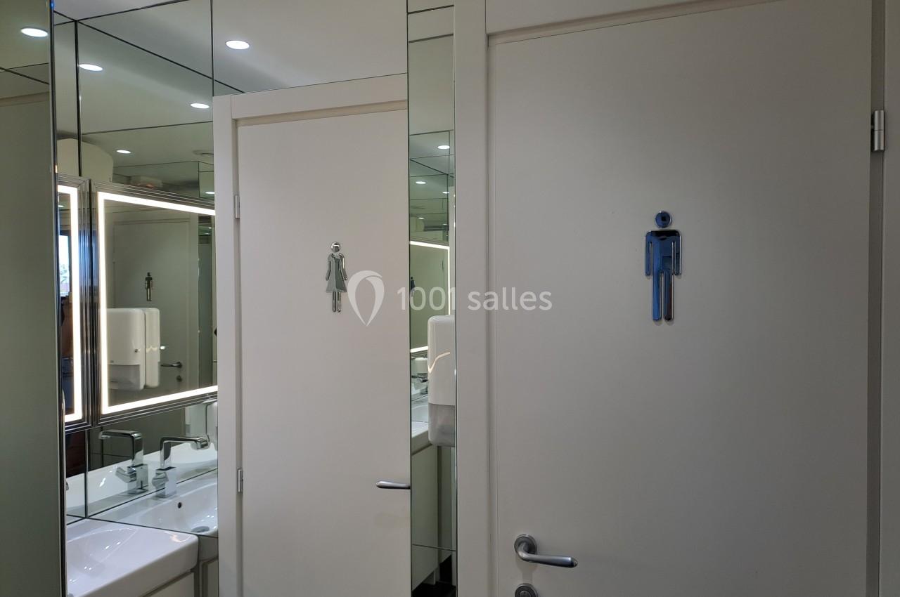 Portes de toilettes avec pictogrammes homme et femme, entourées de miroirs reflétant un lavabo et un éclairage.