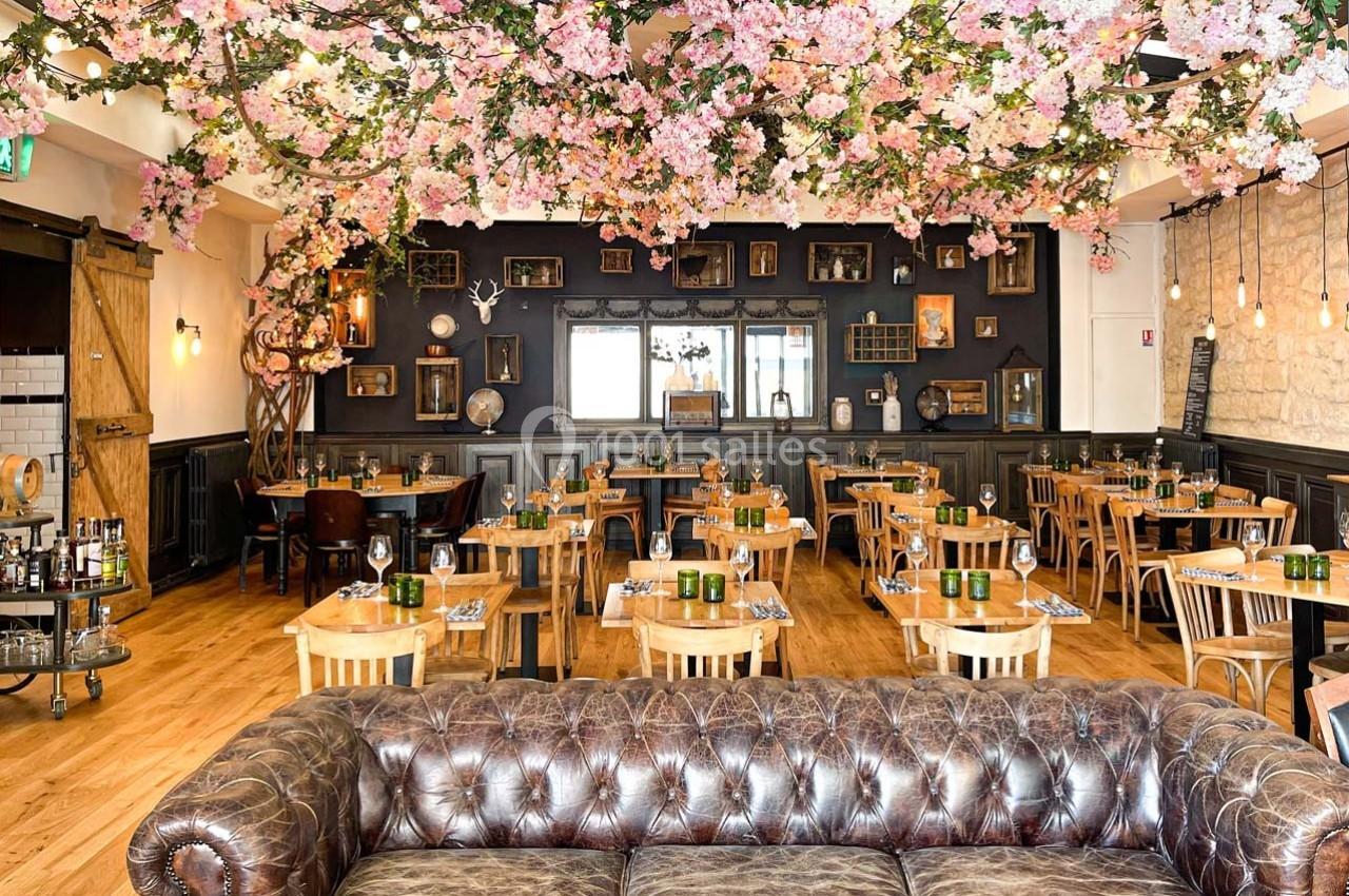 Salle de restaurant décorée de fleurs roses suspendues, avec mobilier en bois et canapé en cuir au premier plan.