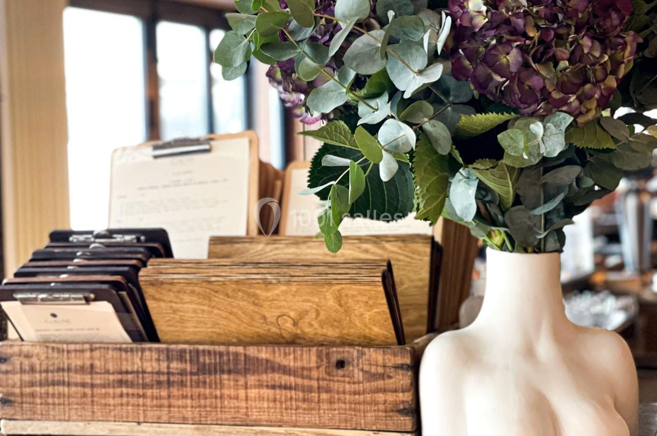 Un vase blanc en forme de buste féminin avec des fleurs violettes et vertes à côté de porte-menus en bois.