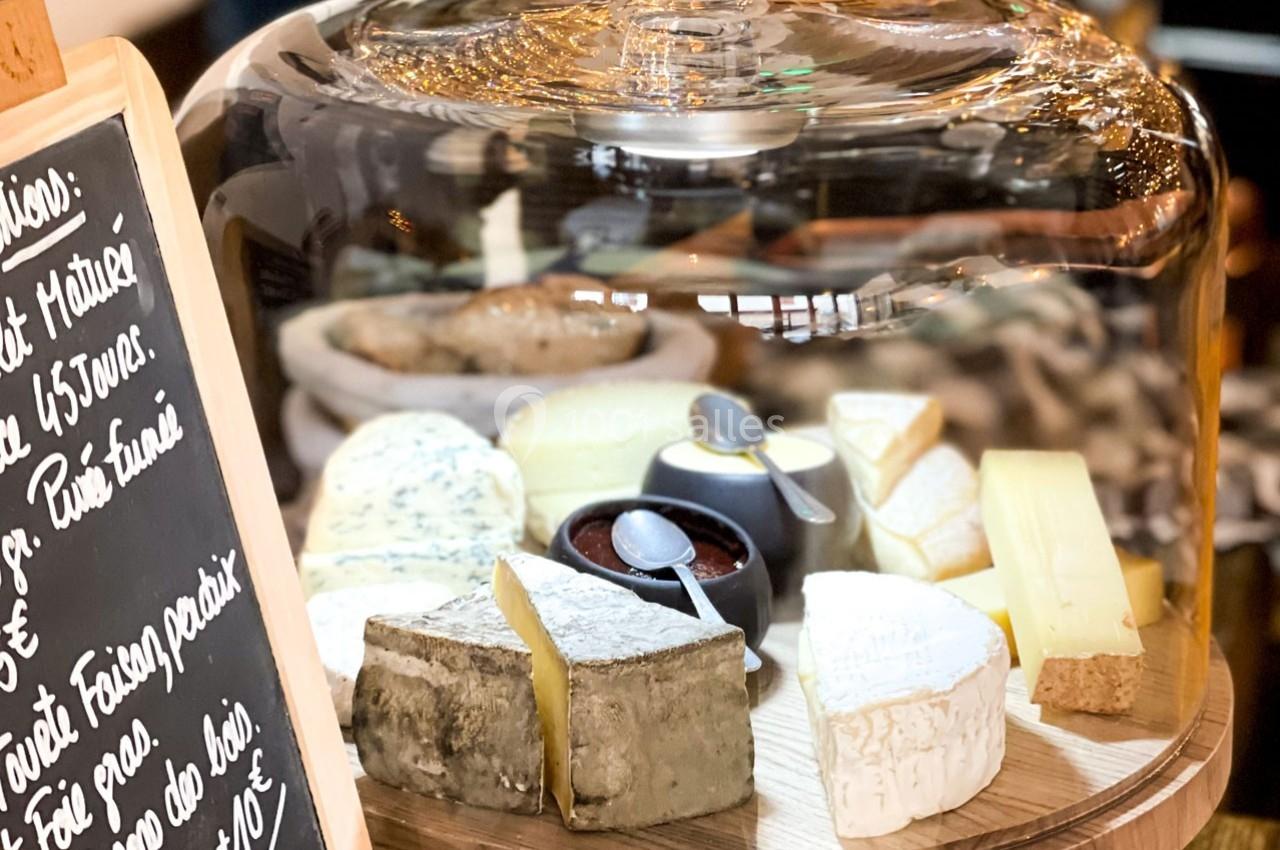 Plateau de fromages variés sous une cloche en verre, accompagné d'un pot de confiture et d'une ardoise avec des inscriptions.