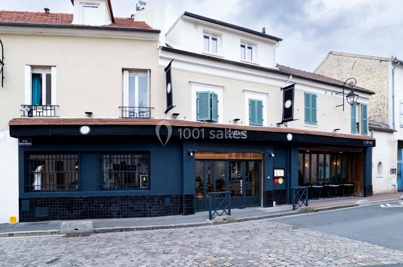 Façade d'un restaurant moderne avec murs bleu foncé, fenêtres vitrées et enseignes noires, situé dans une rue pavée.