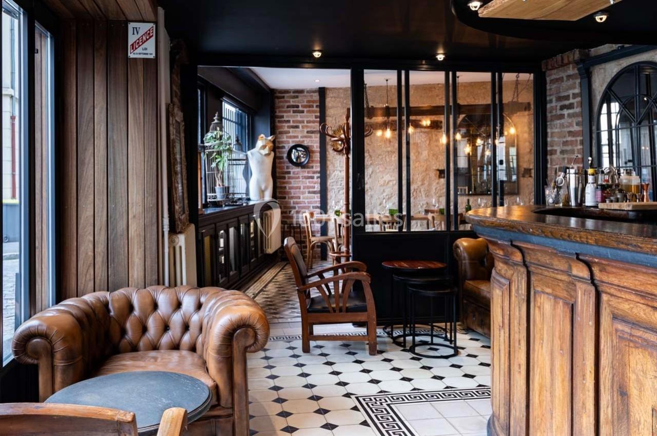 Intérieur d'un café avec fauteuils en cuir, bar en bois, sol en carreaux noirs et blancs, et murs en briques apparentes.