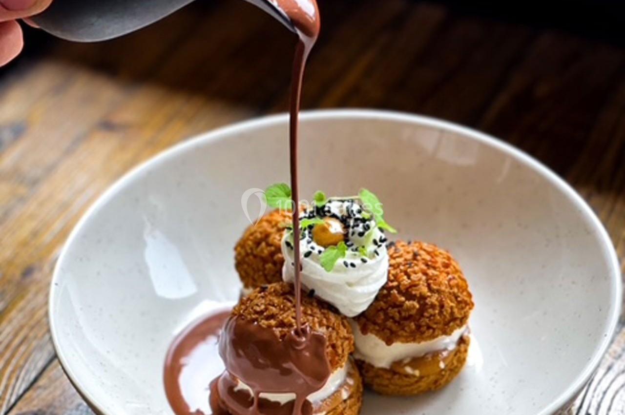 Des choux garnis de crème sont nappés de sauce chocolat, servis dans une assiette blanche sur une table en bois.