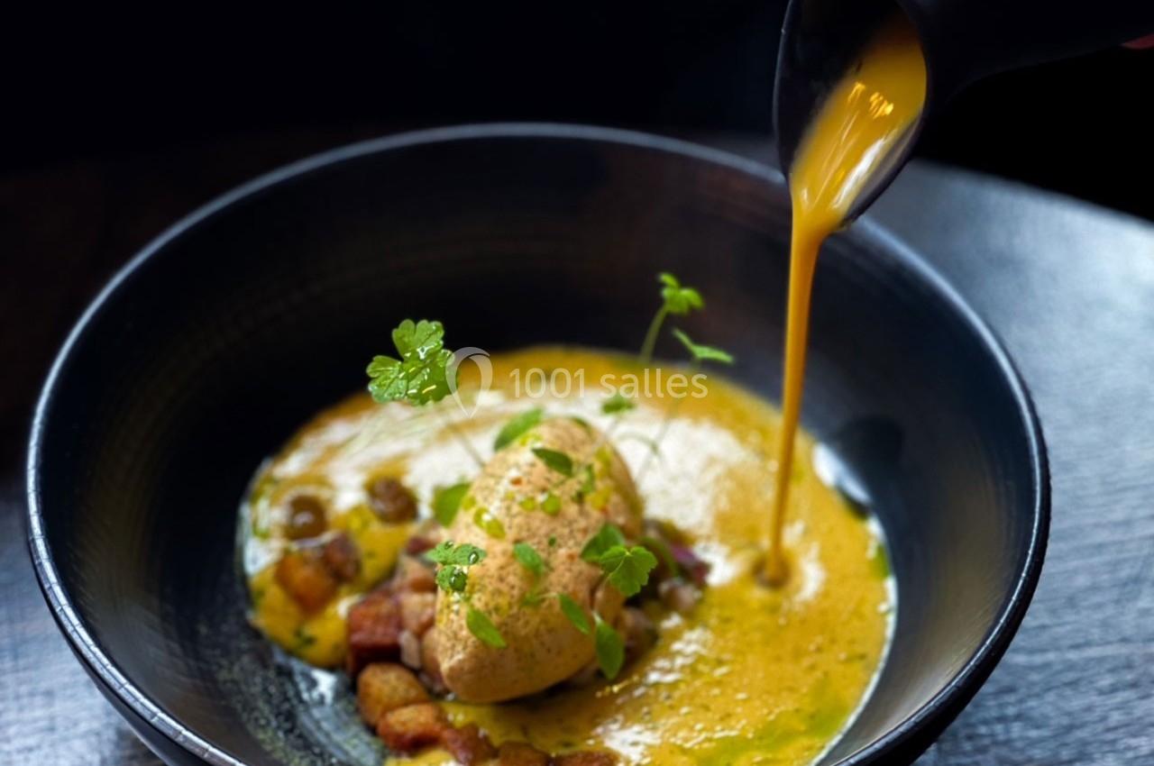 Un velouté orange est versé dans une assiette contenant une quenelle, des croûtons et des herbes fraîches.