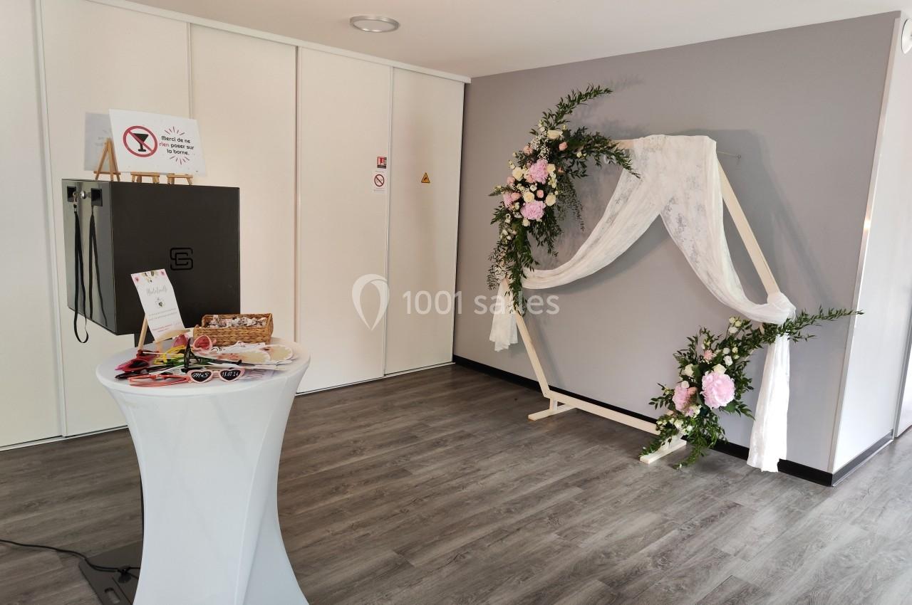 Décoration de salle avec arche florale ornée de voilages blancs et table haute présentant des objets et brochures.