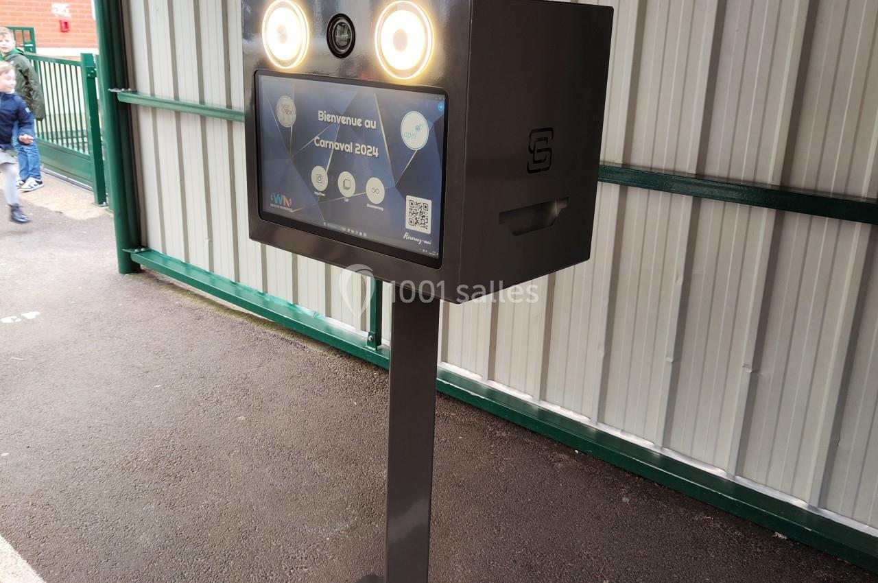 Borne interactive métallique avec deux lumières circulaires, installée sous un abri près d'un mur en tôle ondulée.