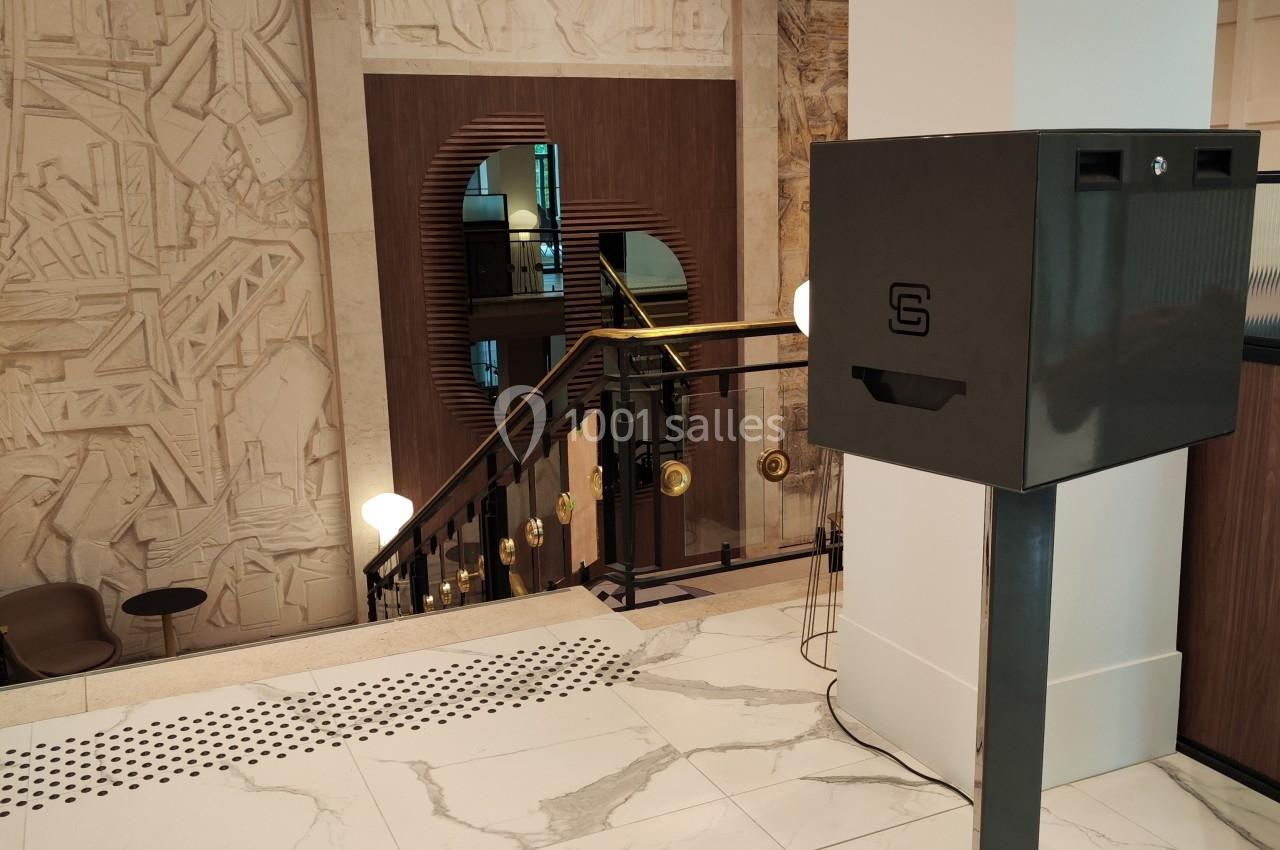 Boîte noire sur pied dans un hall moderne avec escalier, murs ornés de reliefs artistiques et sol en marbre.
