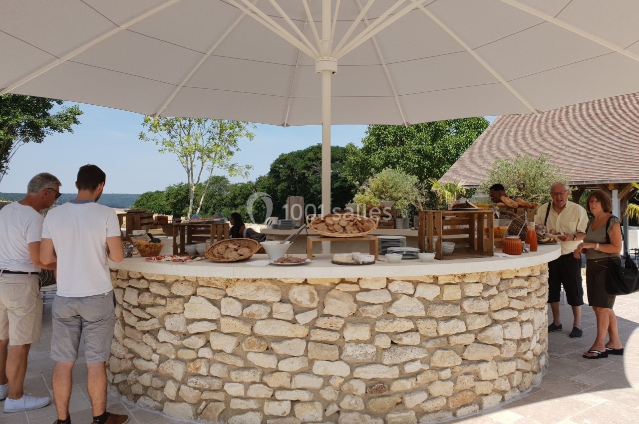 Buffet extérieur sous un grand parasol, avec des plats disposés sur un comptoir en pierre, entouré de convives.