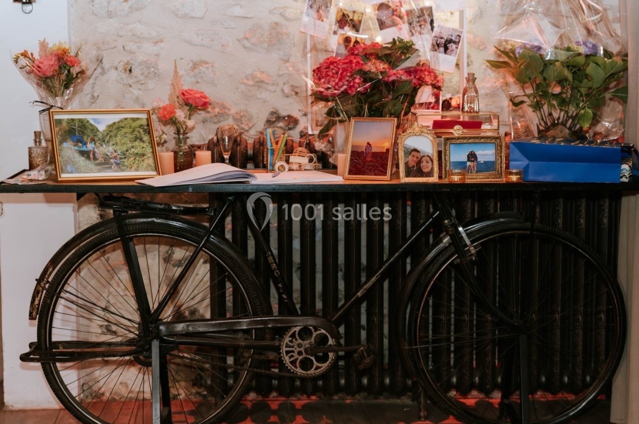 Un vélo ancien appuyé contre un radiateur sous une table décorée de fleurs, cadres photo et objets divers.