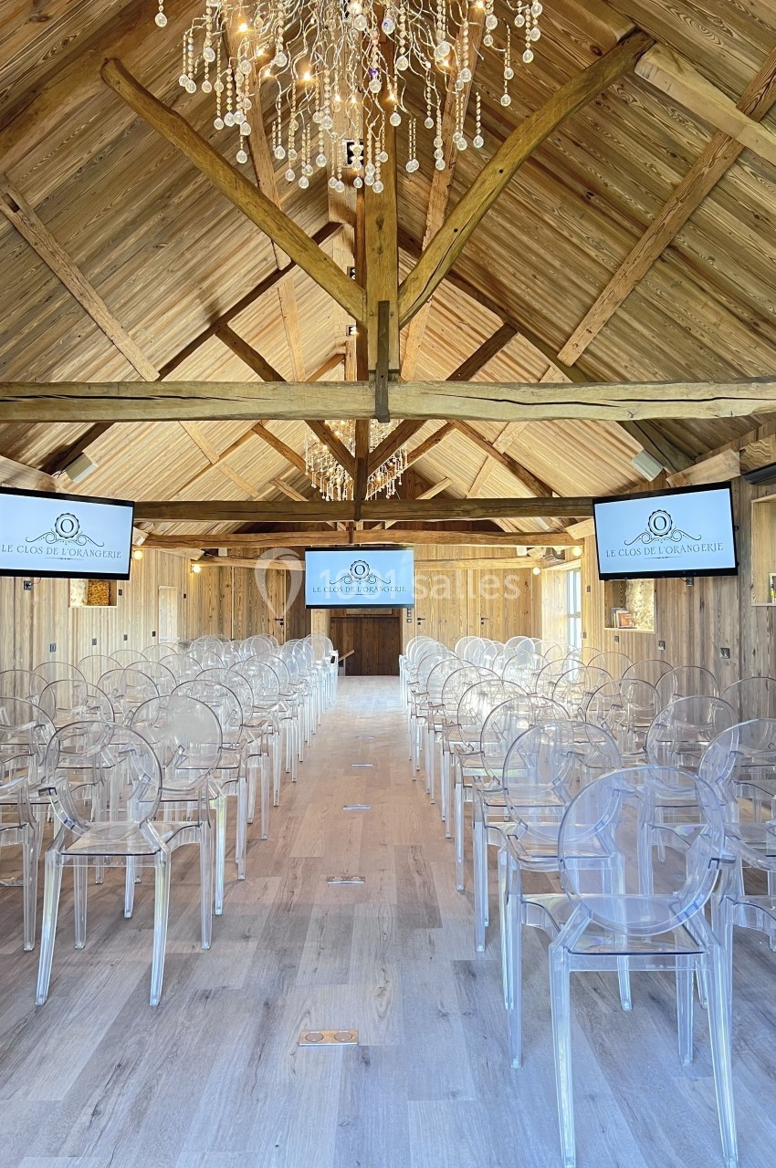 Salle en bois avec plafond en poutres apparentes, rangées de chaises transparentes et écrans affichant un logo.