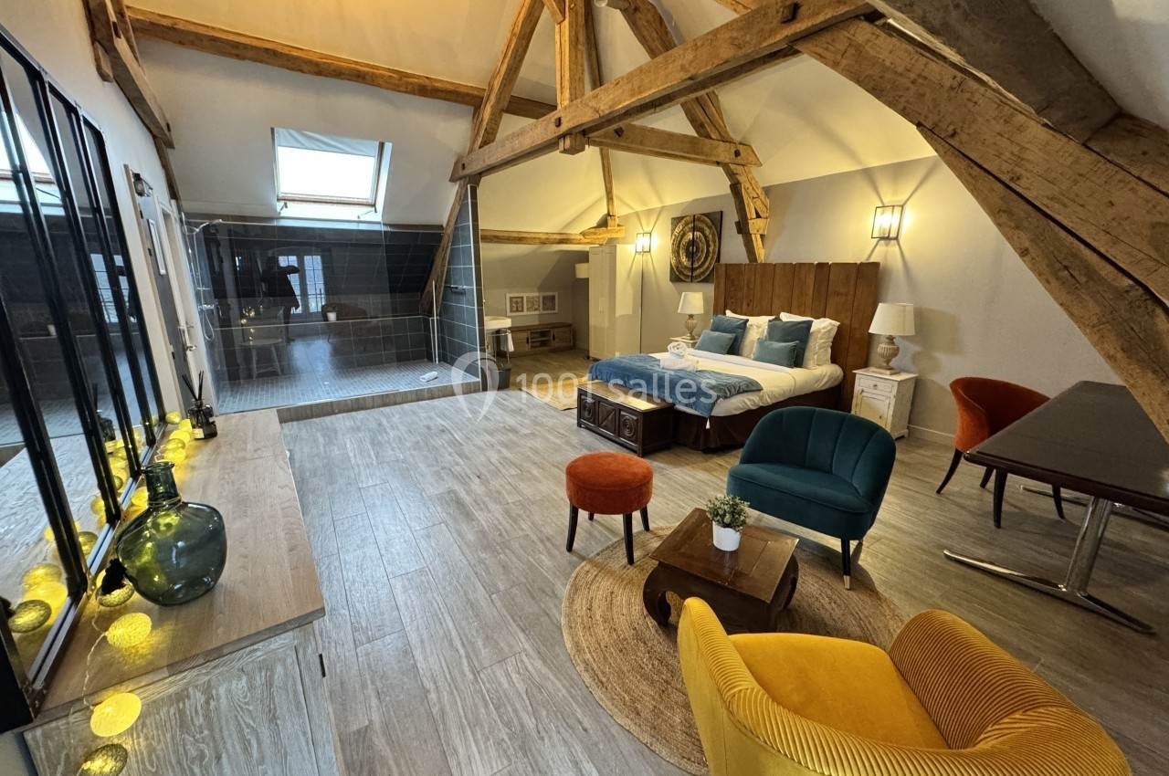 Chambre spacieuse sous combles avec poutres apparentes, lit double, coin salon coloré et grande baie vitrée.