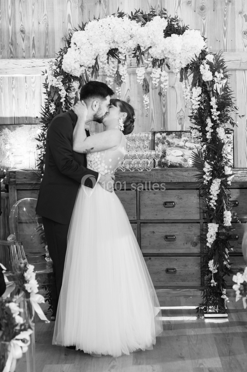 Un couple s'embrasse sous une arche fleurie dans un décor intérieur en bois, lors d'une cérémonie.