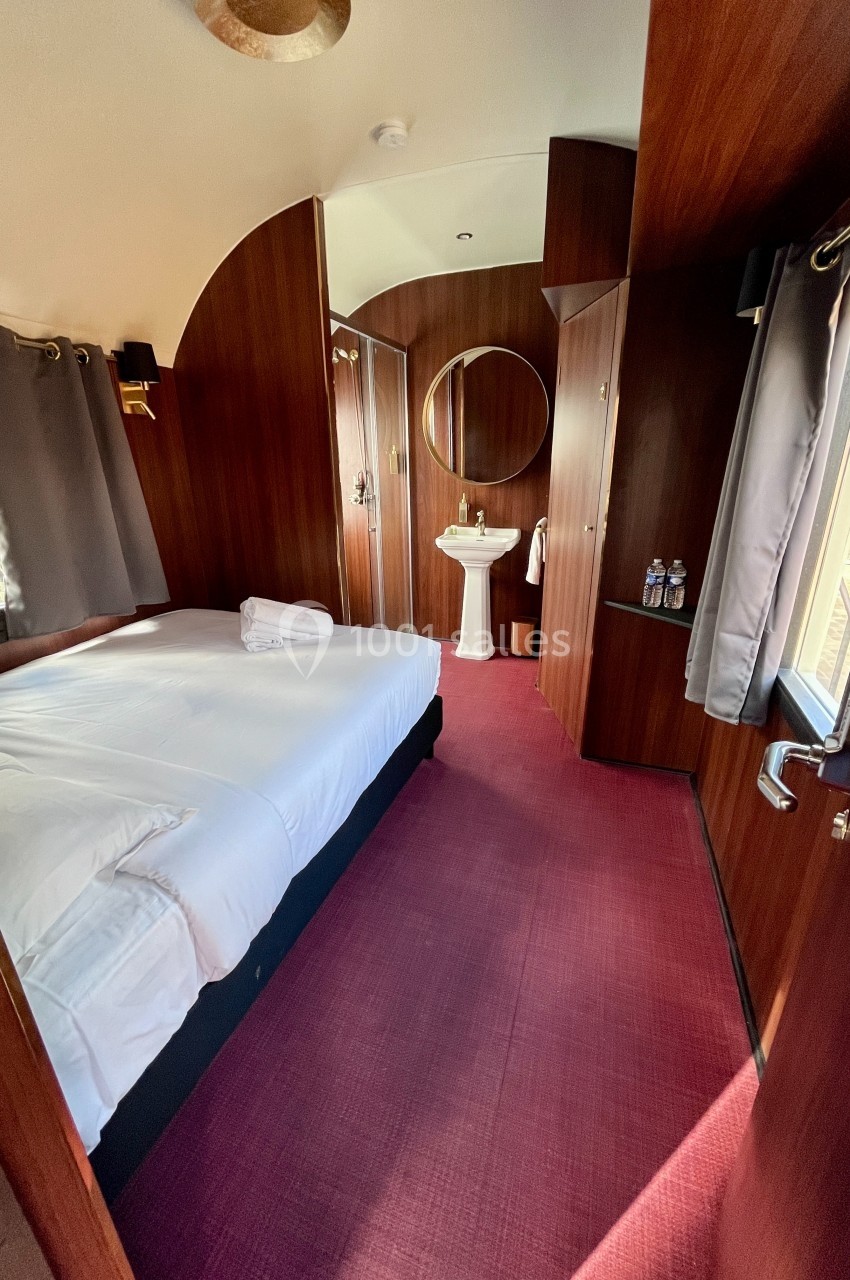 Intérieur d'une chambre avec lit double, rideaux gris, lavabo, miroir rond et sol recouvert de moquette rouge.