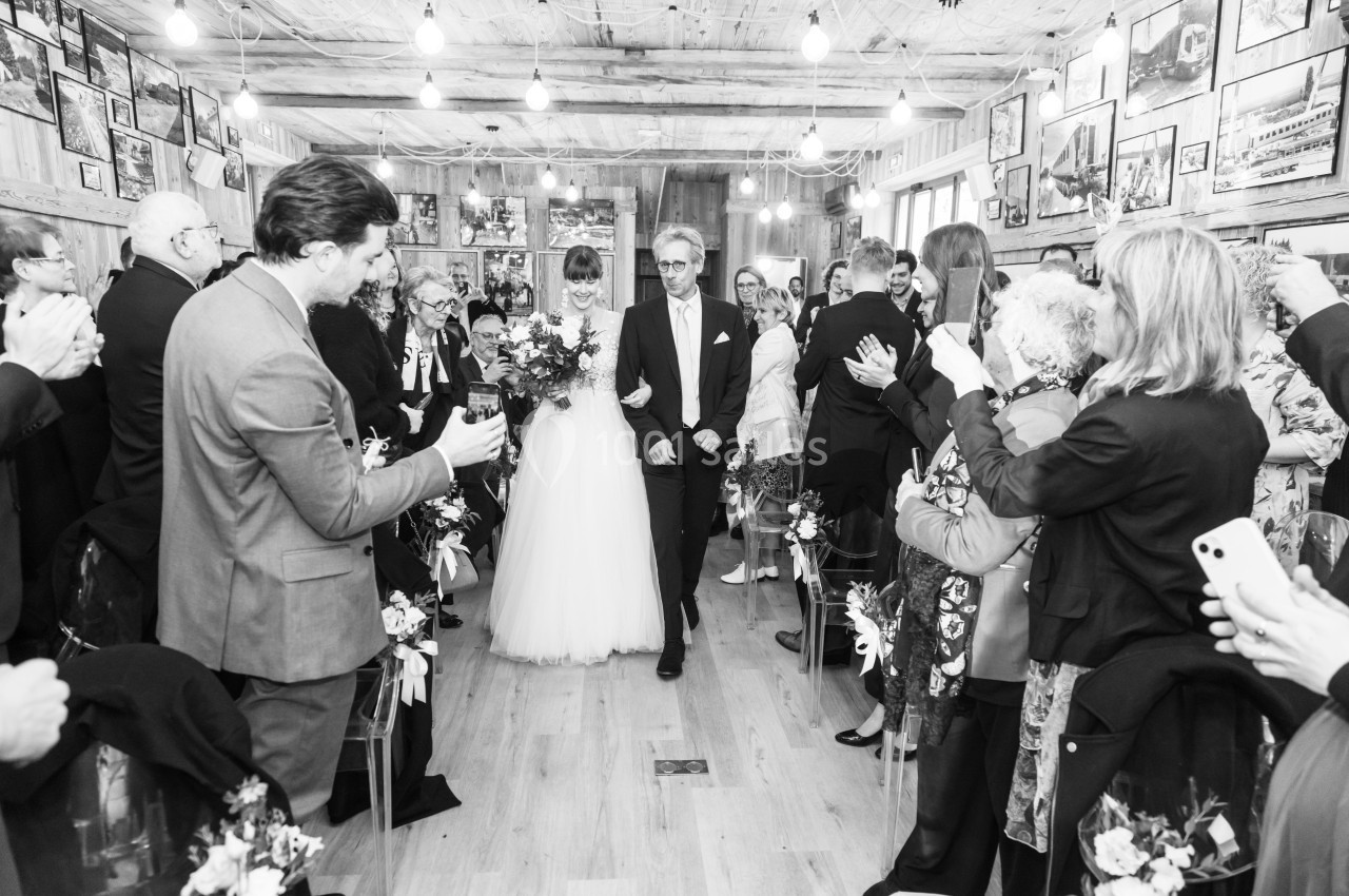 Un couple marche dans l'allée d'une salle décorée pour un mariage, entouré d'invités applaudissant.